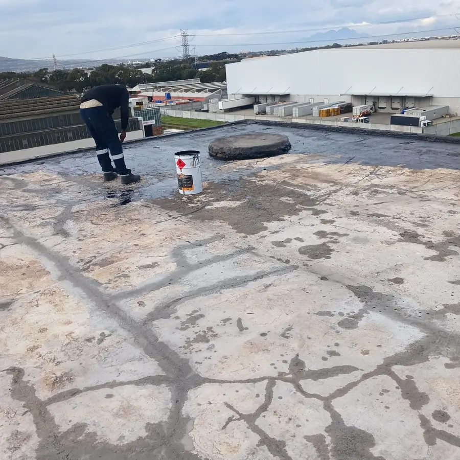 Worker applying bitumen primer to an industrial flat roof before waterproofing